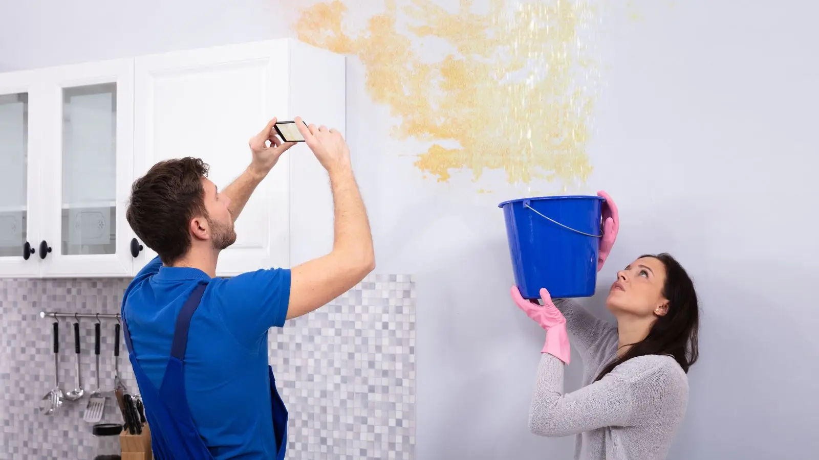 Man photographing a stained wall while a woman holds a bucket beneath the leak